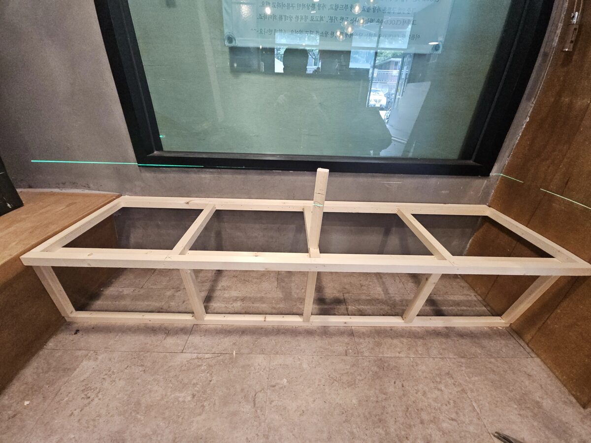 카페 창가에 목재 프레임으로 짜고 있는 벤치형 수납장 골조, 뒤로 유리창에 비친 카페 상호가 보이는 타일 바닥 시공 현장